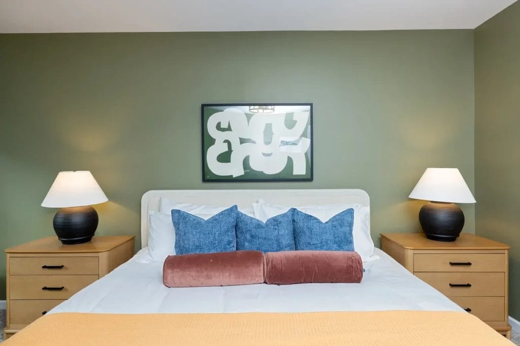 A neatly made bed with blue and pink pillows, flanked by two wooden nightstands with black lamps, sits against an olive green wall featuring an abstract framed artwork.