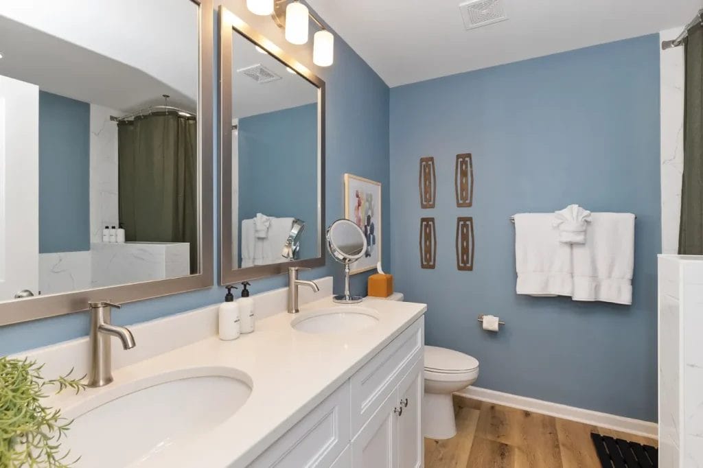 Modern bathroom with blue walls, double sink vanity, large mirror, wooden floor, toilet, towel with a swan-shaped fold, framed art, and a shower with a dark green curtain in the background.