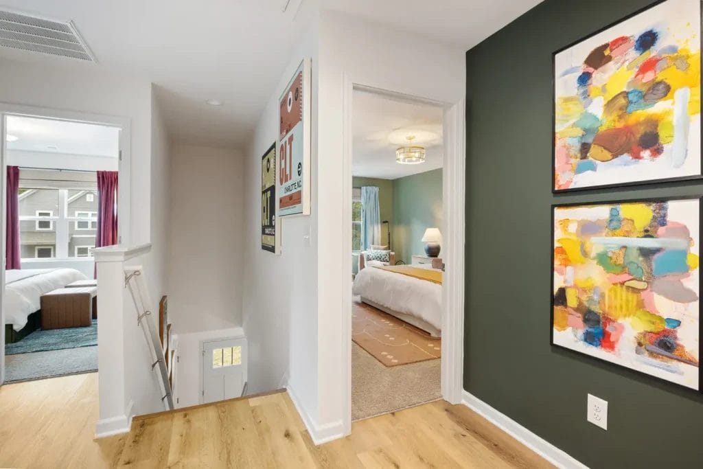 A bright hallway with light wood floors, colorful abstract art on a dark green wall, and doors leading to two bedrooms. One bedroom has a bed with white linens; another bed with dark bedding is visible to the left.