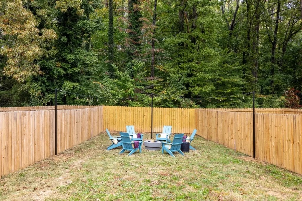 A fenced backyard area with six blue Adirondack chairs arranged in a circle around a fire pit, surrounded by lush green trees and grass.
