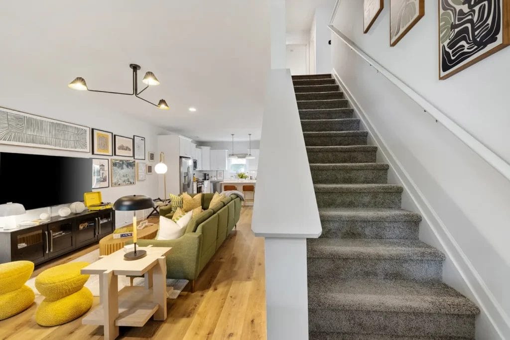 A modern living room with a green sofa, yellow ottomans, wall art, and a flat-screen TV. Wooden floors and a staircase with gray carpet lead to the upper floor. The open kitchen is visible in the background.