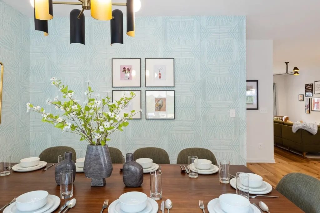 A modern dining room with a wooden table set for six, featuring white dishes and glassware. A vase with white flowers and smaller vases are centered on the table. Geometric wallpaper and framed art decorate the background wall.