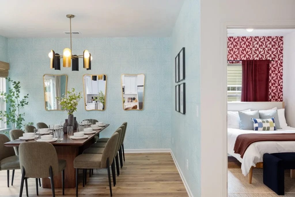 Modern dining room with a wooden table set for eight, green chairs, three gold-framed mirrors on a patterned wall, and a view into a bedroom with a red accent wall and neatly made bed.