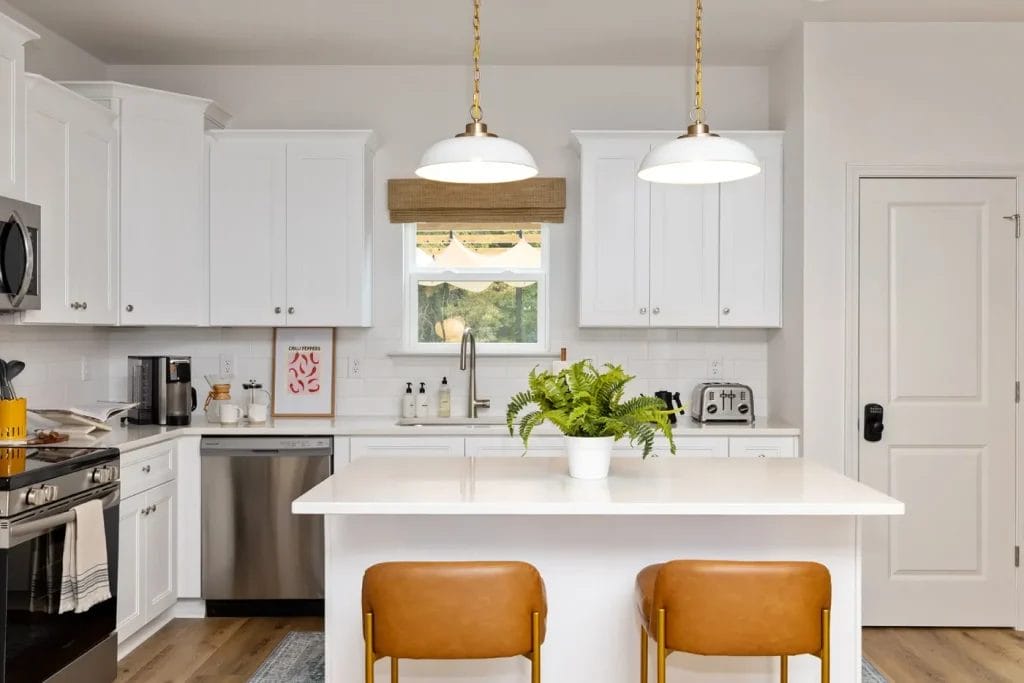 Bright modern kitchen with white cabinets, a center island with a white countertop, two tan leather stools, pendant lights, stainless steel appliances, a window above the sink, and a potted plant on the island.