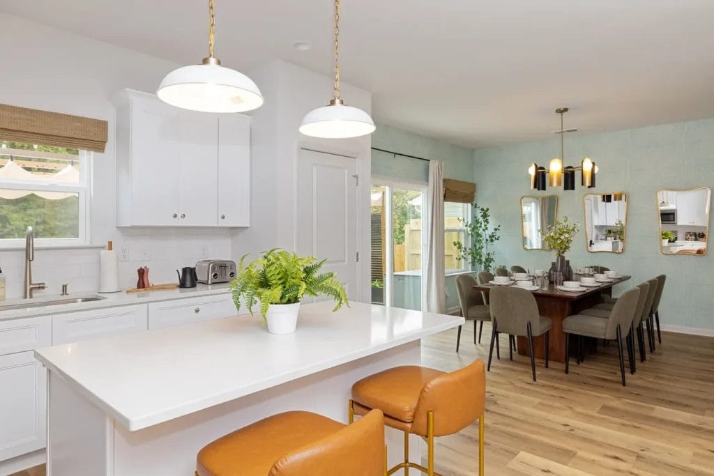 Modern kitchen and dining area with white cabinets, a large island with two brown barstools, a potted plant, wooden flooring, and a dining table set for a meal under contemporary lighting.