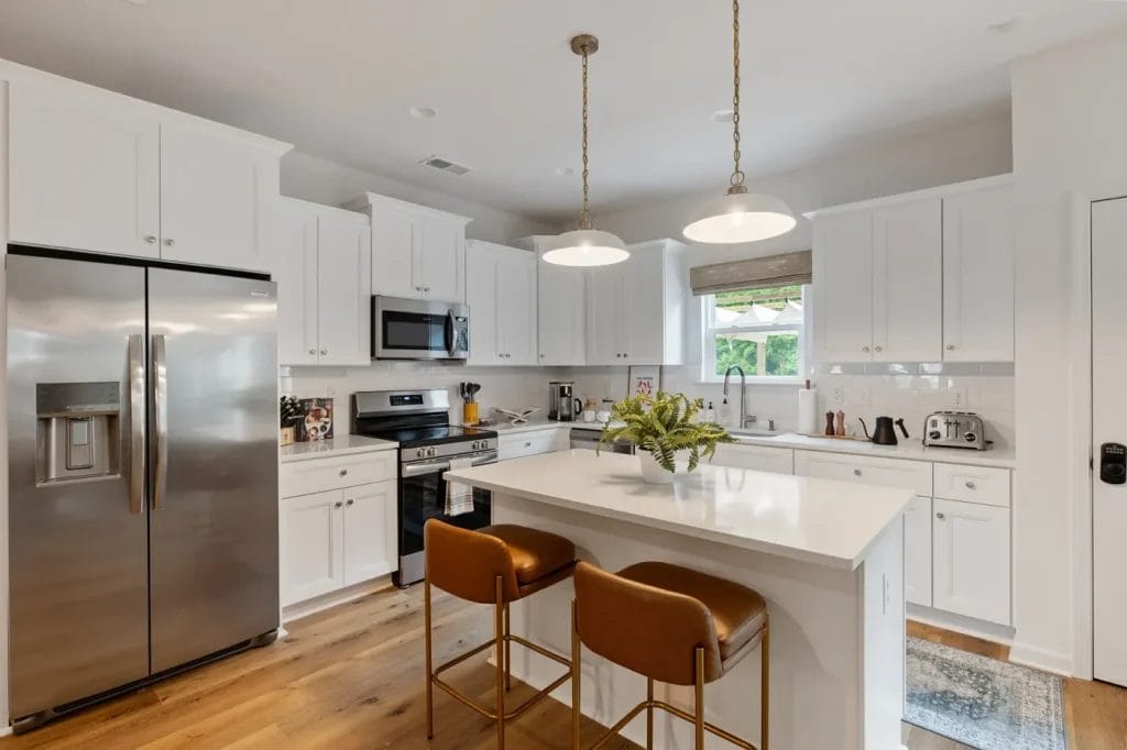 Modern white kitchen with stainless steel appliances, an island with two brown leather bar stools, pendant lights above, wooden floors, and a window over the sink letting in natural light.