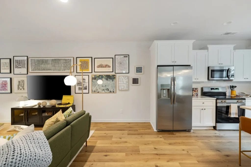 A modern living room with a green sofa, wall-mounted TV, and a gallery wall of framed art, adjacent to a kitchen with white cabinets, stainless steel appliances, and light wood flooring.