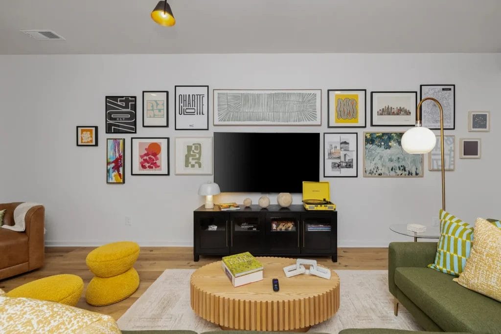 Modern living room with a gallery wall of framed art above a black TV stand, TV, and decor items. There's a round wood coffee table, green sofa with yellow pillows, yellow stools, and a floor lamp, all on a light rug.