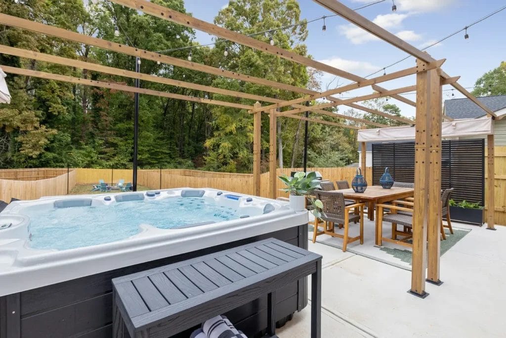 A backyard patio with a pergola, featuring a bubbling hot tub, a bench with towels, a dining table set for six, potted plants, and string lights overhead. Trees and a wooden fence border the area.