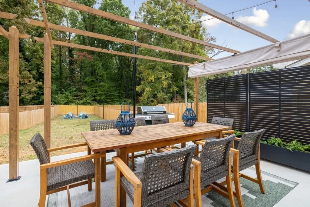 A wooden outdoor dining table with six woven chairs sits under a pergola on a patio. Two blue lanterns are on the table. Behind, there’s a grill, privacy screen, yard, and trees.