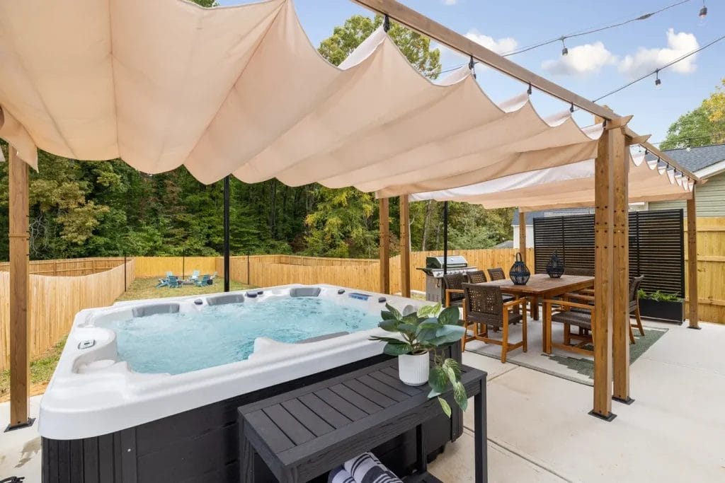 A backyard patio with a hot tub under a canopy, next to an outdoor dining table and chairs. String lights are hung above, and there is a potted plant on a bench. A wooden fence and trees are in the background.