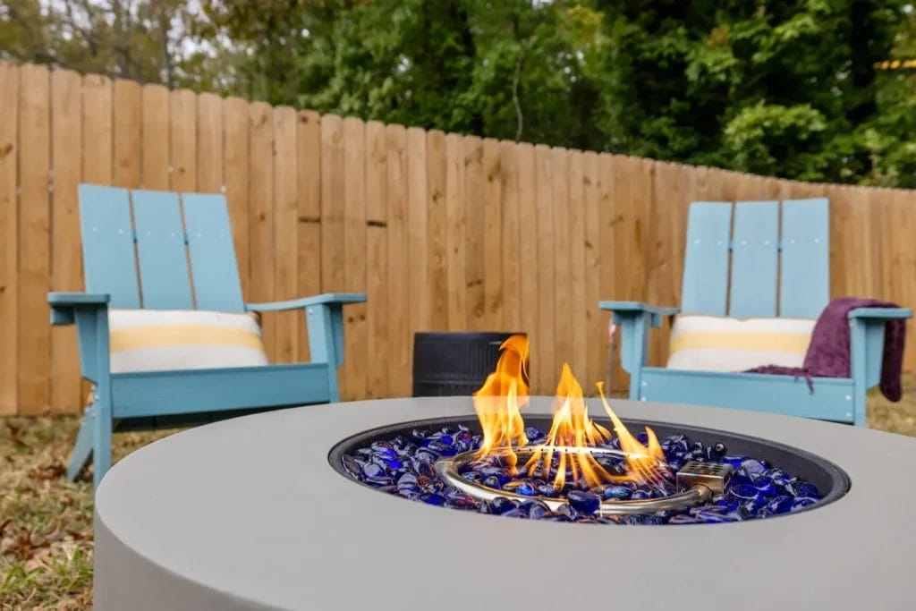A modern outdoor fire pit with blue glass rocks burning in the foreground, flanked by two blue wooden Adirondack chairs with white cushions, set on a grassy lawn near a wooden privacy fence.