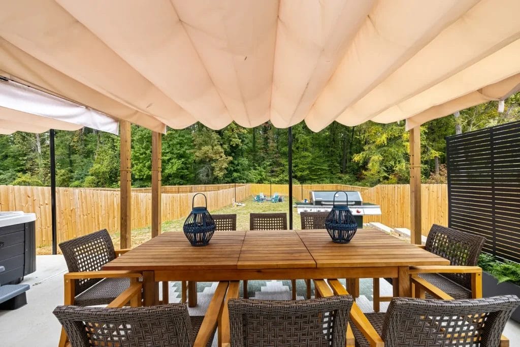 A covered outdoor patio features a wooden dining table with eight woven chairs, two decorative lanterns on the table, a grill in the background, and a fenced yard surrounded by trees.