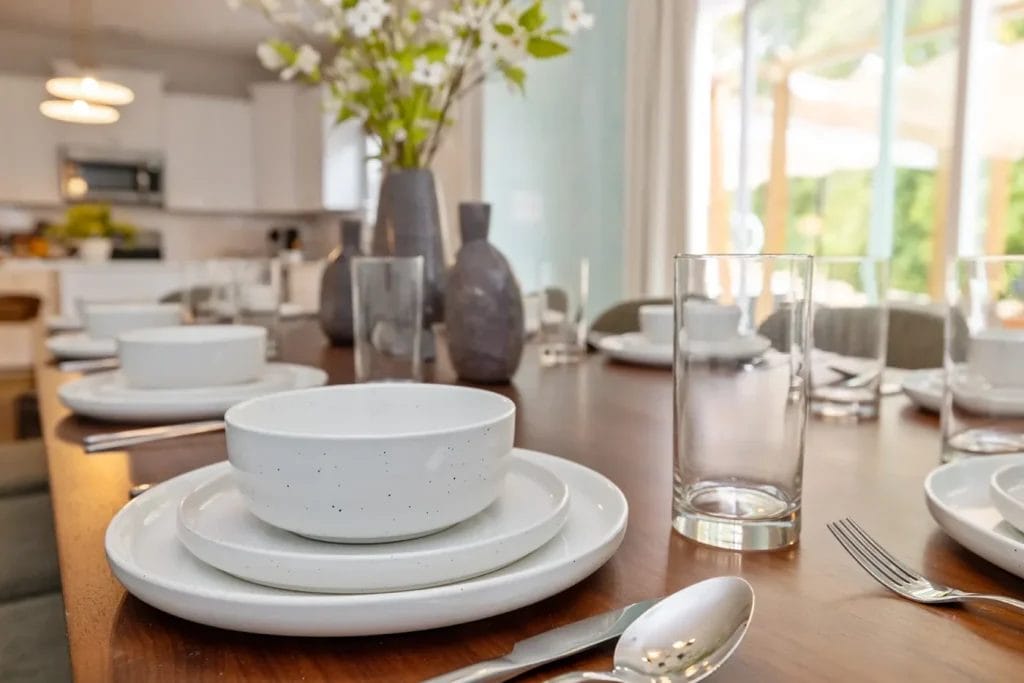 Close-up of a wooden dining table set with white dinnerware, glasses, silverware, and decorative vases with flowers. A bright, modern kitchen and large windows with greenery outside are visible in the background.