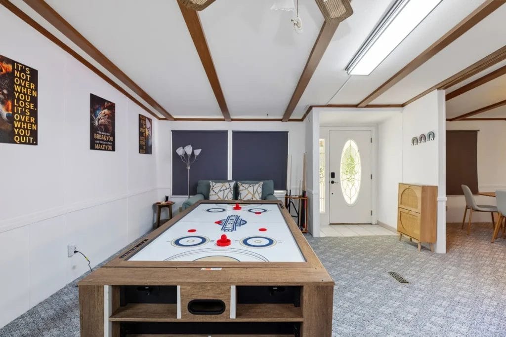 A living room with an air hockey table in the center, a sofa against the back wall, movie posters and motivational quotes on the walls, and a dining area visible to the right through an open doorway.