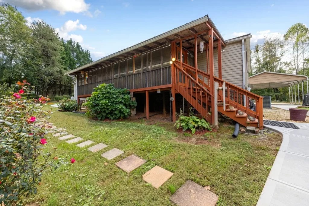 A wooden house with a covered, screened-in porch and stairs, surrounded by green grass, plants, and stepping stones. A carport is visible to the right, and trees are in the background under a partly cloudy sky.