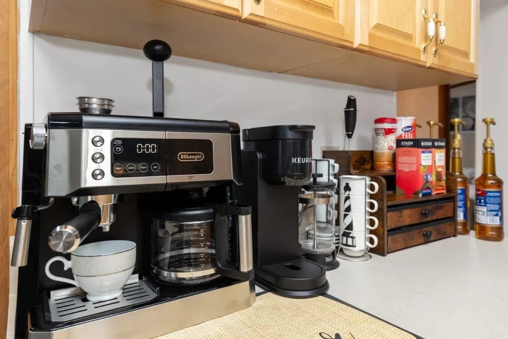 A kitchen countertop with a De'Longhi espresso machine, a Keurig coffee maker, a cup, a mug holder, coffee supplies, and several syrup bottles next to wooden cabinets.