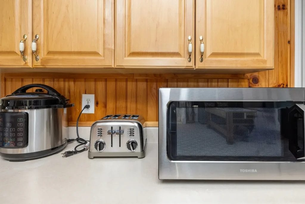 A kitchen countertop with a pressure cooker, a stainless steel toaster, and a Toshiba microwave oven, set against wooden cabinets and a wood-paneled backsplash.
