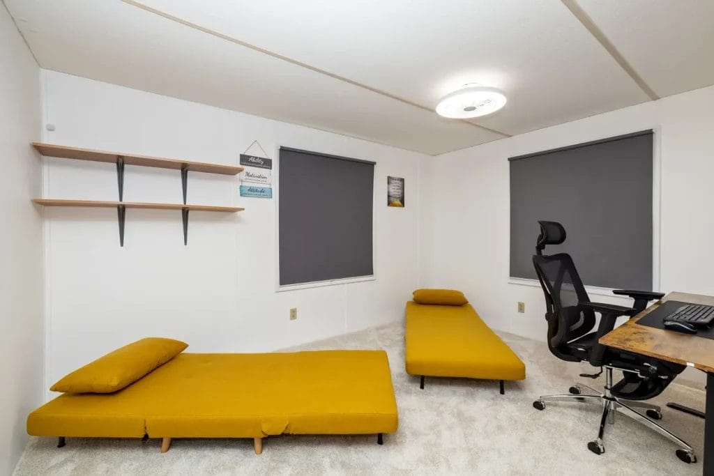 A minimalist room with white walls, two mustard yellow floor sofas, a black office chair, a wooden desk, wall shelves, and two windows with dark blinds. The carpet is light-colored.