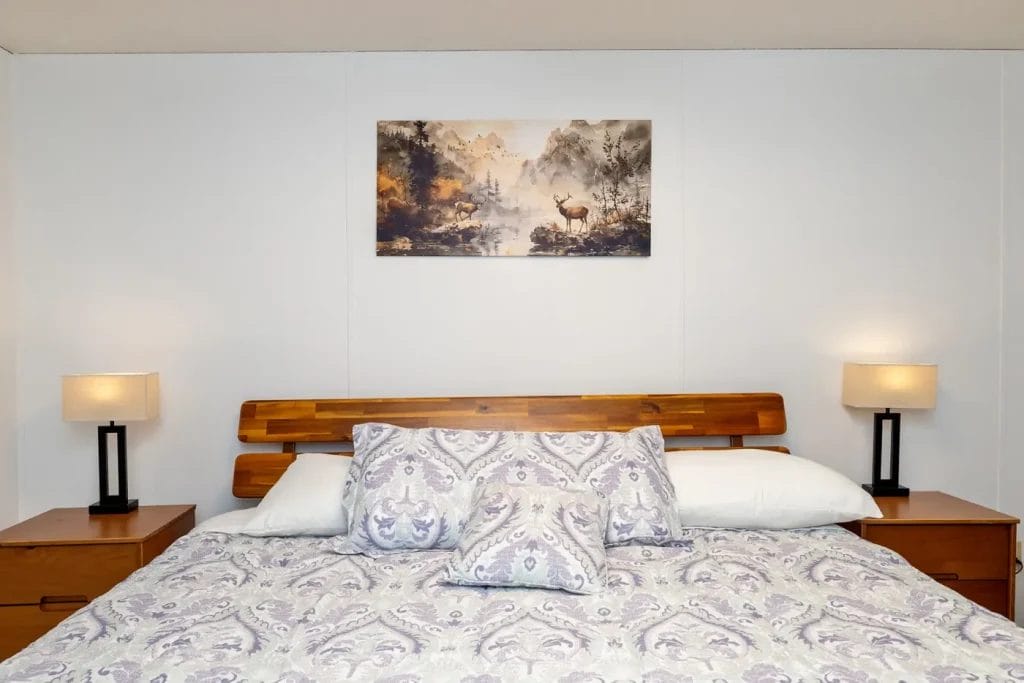 A cozy bedroom with a wooden headboard, patterned bedding, two pillows, and matching nightstands with lamps. A nature-themed artwork featuring a deer hangs on the white wall above the bed.