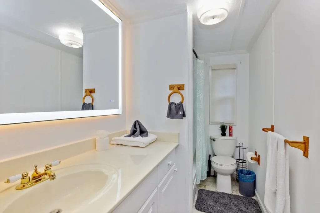 A clean, white bathroom with a large illuminated mirror above a sink, gold faucet, towels on wooden racks, a toilet with accessories nearby, and a shower with a light blue curtain.