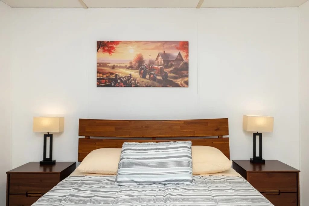 A neatly made bed with striped bedding, two beige pillows, and a wooden headboard sits between two nightstands with square lamps. Above the bed hangs a painting of a rural scene with a tractor and farm buildings.