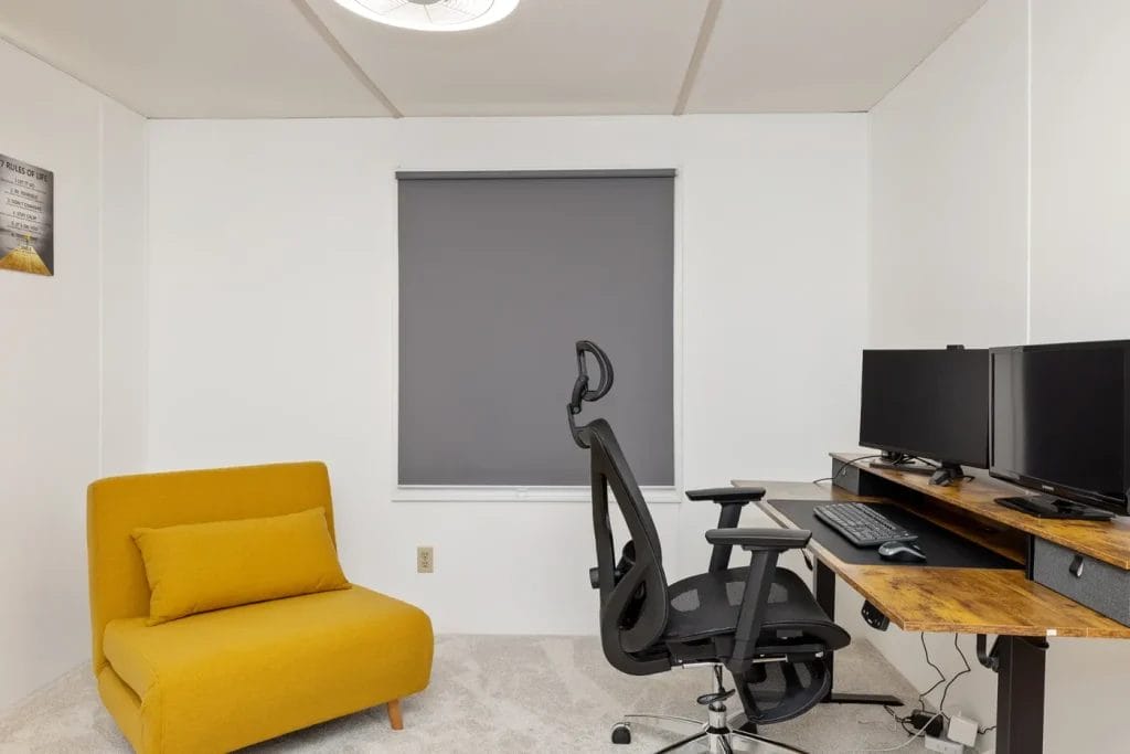 A minimalist office with a yellow lounge chair, a black ergonomic office chair at a wooden desk with dual monitors, keyboard, and mouse. The window behind the desk has a closed gray roller shade.