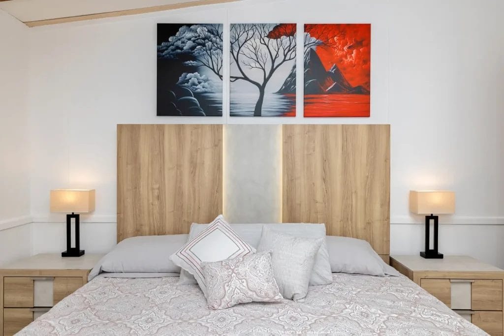 A modern bedroom with a large wooden headboard, two bedside tables with lamps, a neatly made bed with patterned bedding and pillows, and a three-piece wall art featuring abstract trees and mountains above the bed.