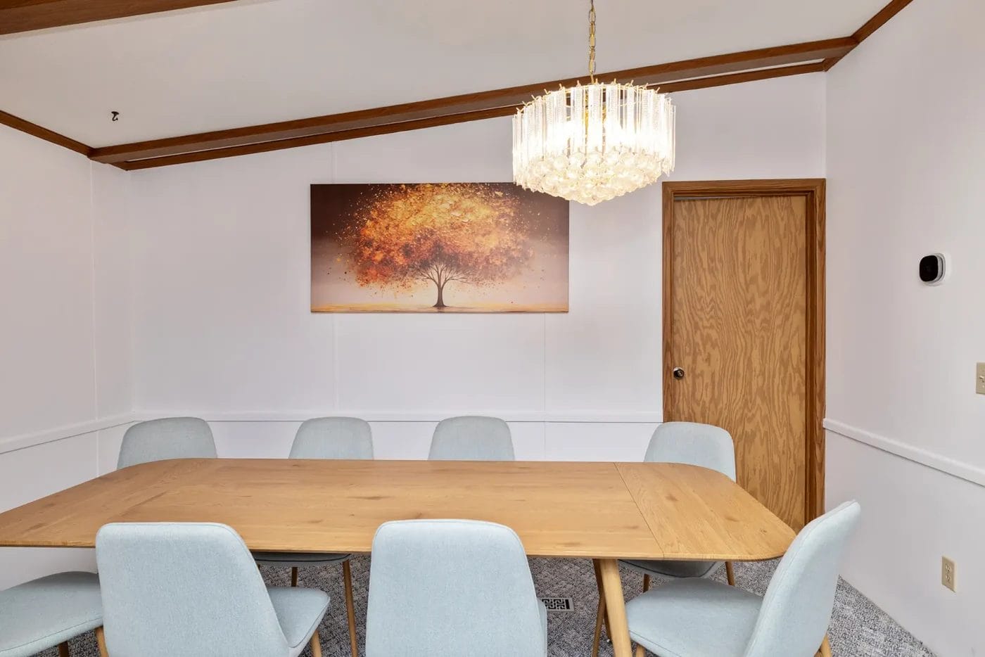 A modern dining room with a wooden table, six light gray chairs, a crystal chandelier, and a wall painting of a tree with golden leaves above a wooden door.