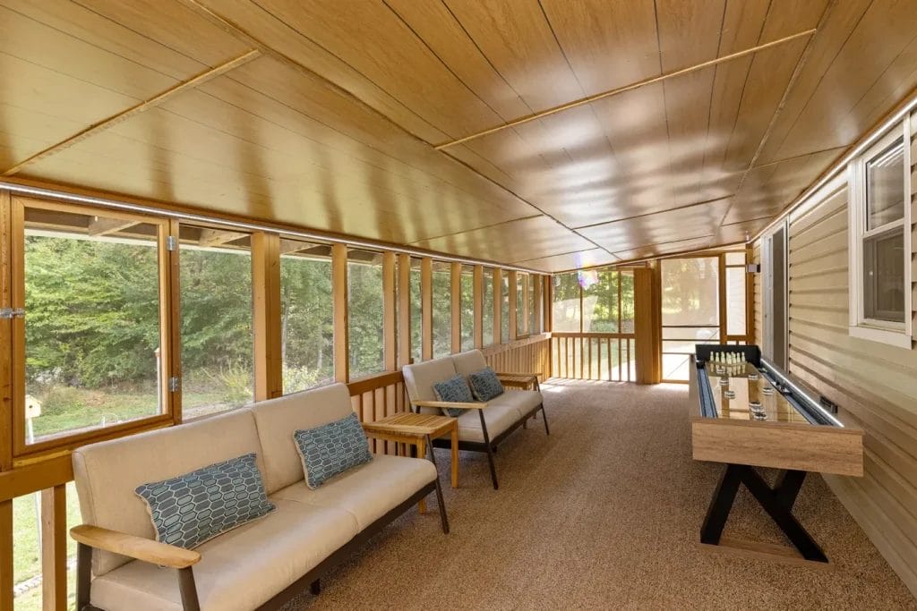 A long, enclosed sunroom with wood-paneled ceiling and walls, large windows overlooking greenery, beige cushioned seating, blue accent pillows, and a wooden console table. Natural light fills the space.
