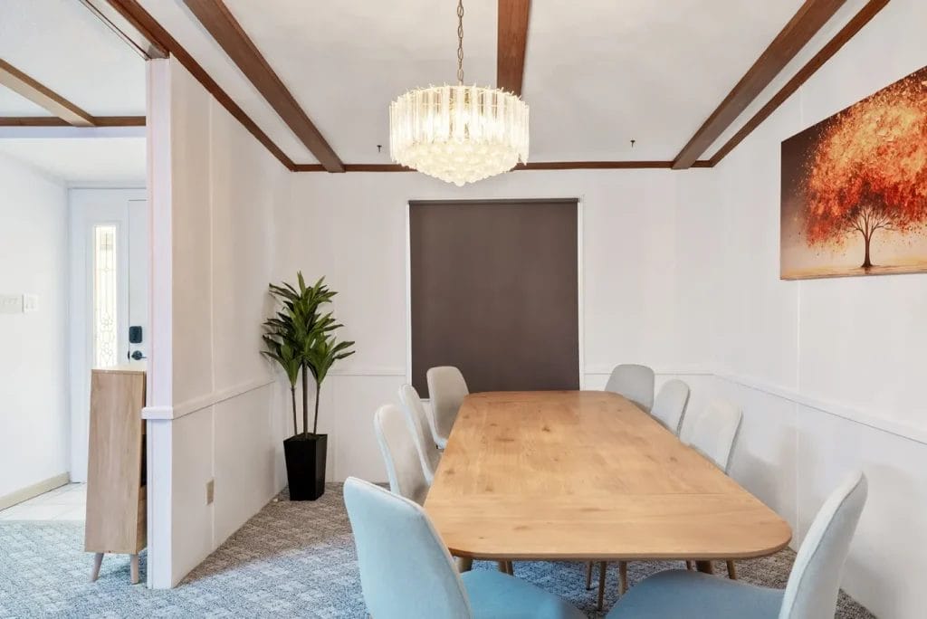 A modern dining room with a wooden table, eight light blue chairs, a potted plant, a crystal chandelier, and a wall painting of an orange tree. The room has white walls and exposed wooden ceiling beams.