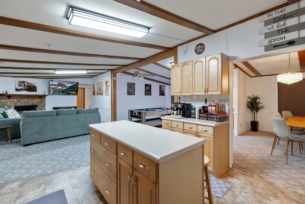 A spacious open-concept kitchen with light wood cabinets and an island, leading to a living area with sofas and a fireplace. A dining area with a modern table and chandelier is visible in the background.