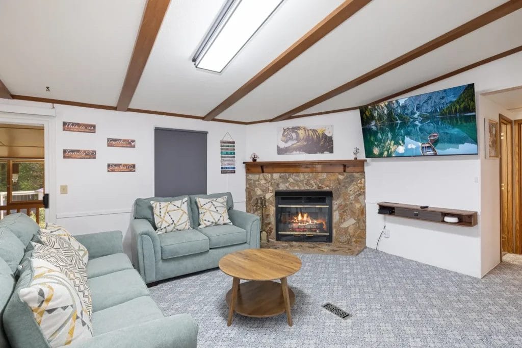 Cozy living room with two blue sofas, a round wooden coffee table, and a fireplace below a wall-mounted TV. The room has wood ceiling beams, patterned carpet, and wall art featuring nature and inspirational quotes.