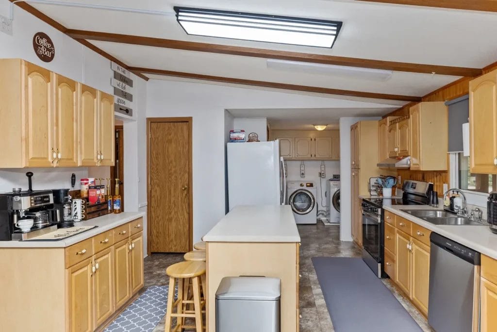 A kitchen with light wood cabinets, an island with two stools, stainless steel appliances, and a coffee station. A refrigerator and a laundry area with washer and dryer are visible in the background.