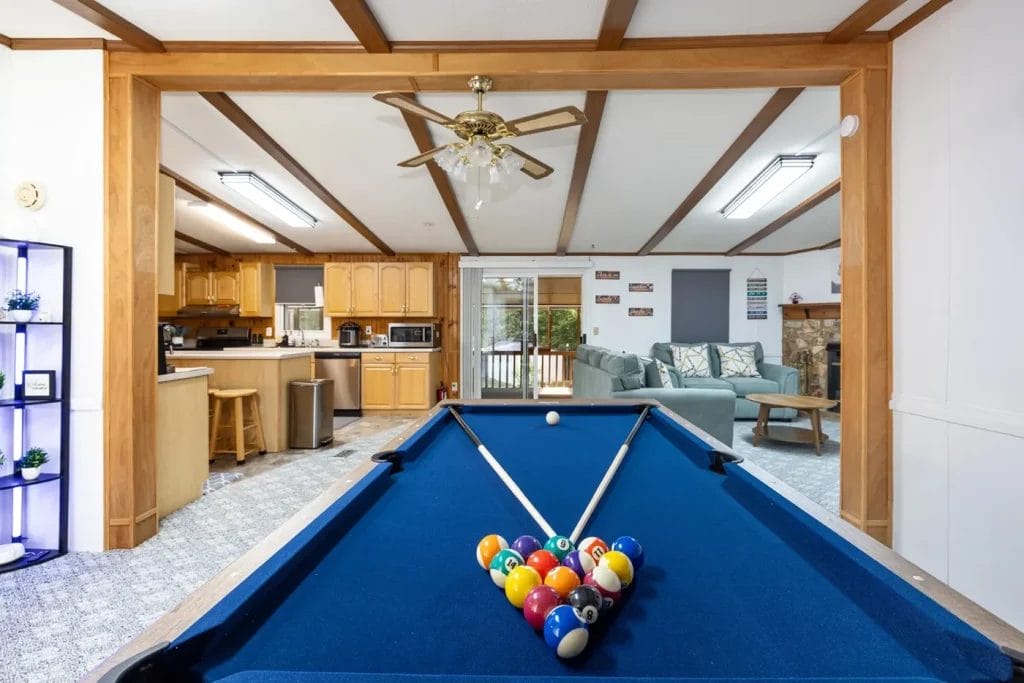 A blue pool table with racked balls sits in a spacious, open living area with a kitchen, ceiling fan, sectional sofa, fireplace, and large windows letting in natural light.
