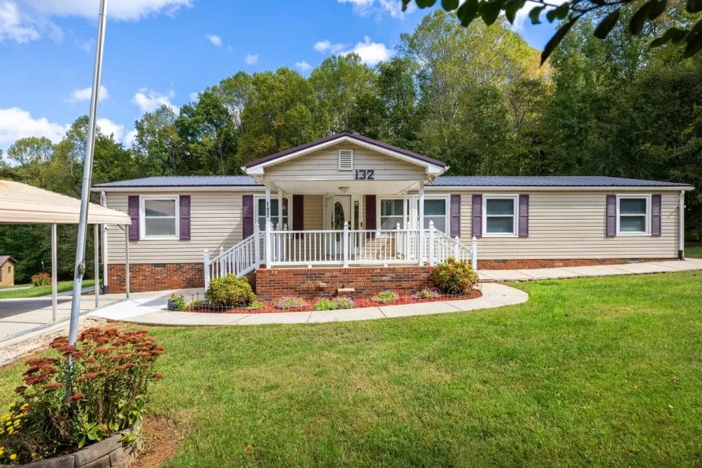 A single-story beige house with purple shutters, a brick foundation, and a white front porch with railings, surrounded by green grass, shrubs, and trees under a blue sky with clouds.