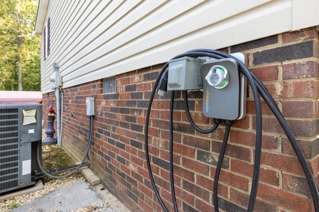 An electric vehicle charging station with cables is mounted on the brick exterior wall of a house, next to an air conditioning unit and other electrical boxes.