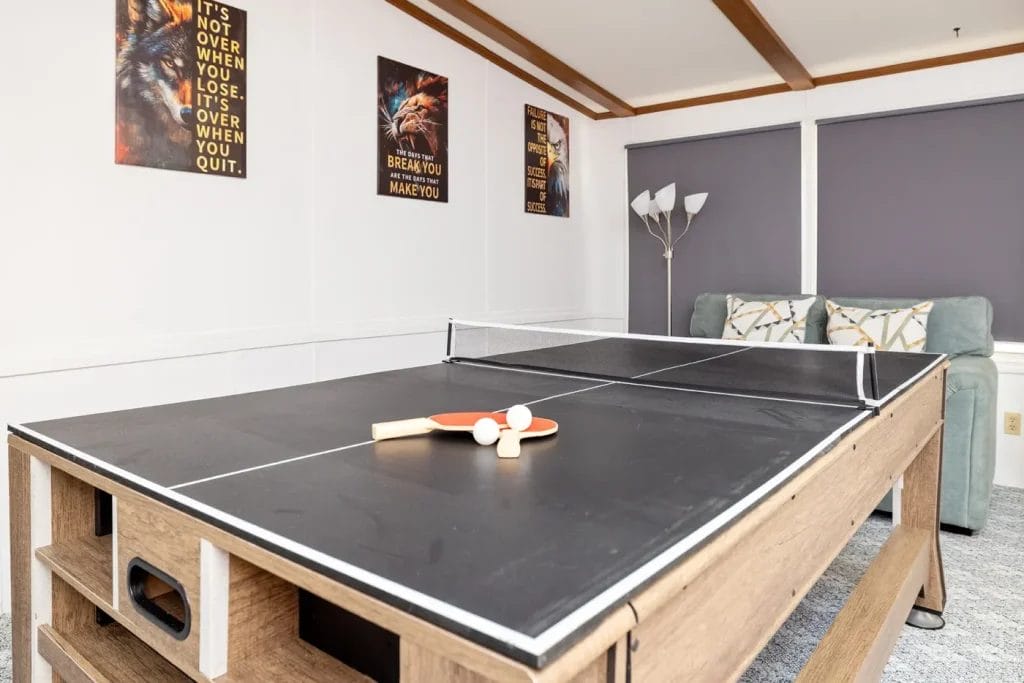 A black table tennis table with paddles and balls sits in a bright room with a gray couch, floor lamp, and motivational posters on the walls.