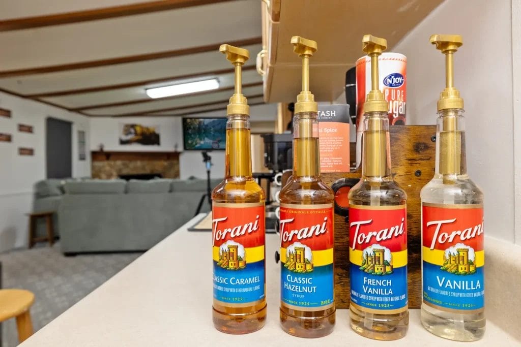Four Torani syrup bottles with pumps—Classic Caramel, Classic Hazelnut, French Vanilla, and Vanilla—are lined up on a counter in a cozy living room with a couch and fireplace in the background.