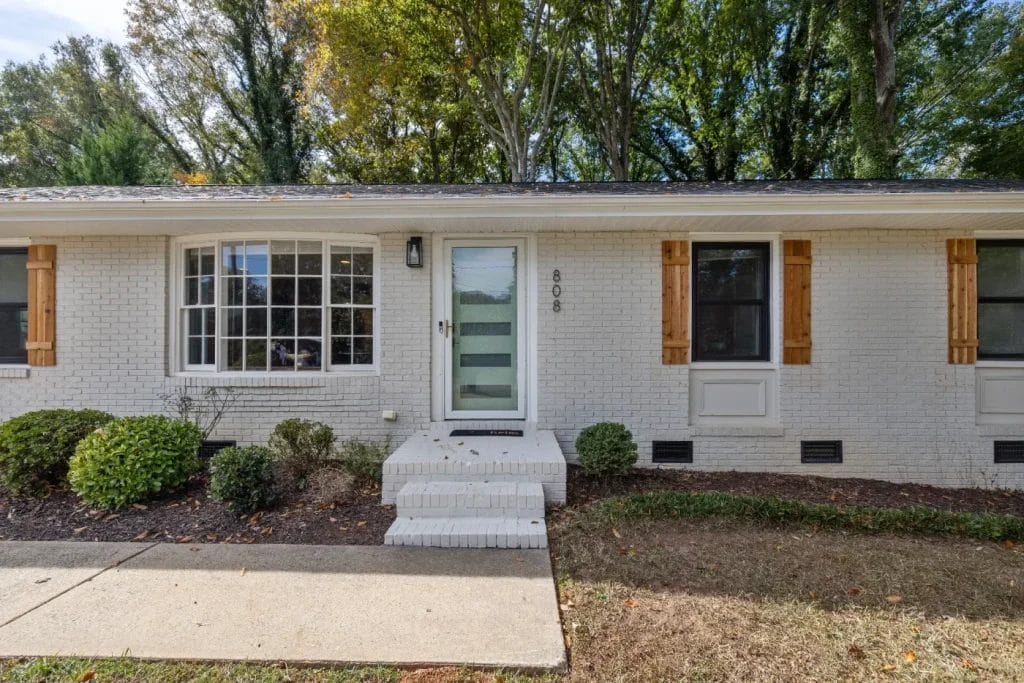 A white brick house with a modern glass front door, flanked by two windows with wooden shutters. There are small shrubs in front and a concrete walkway leading to the entrance. Tall trees are in the background.