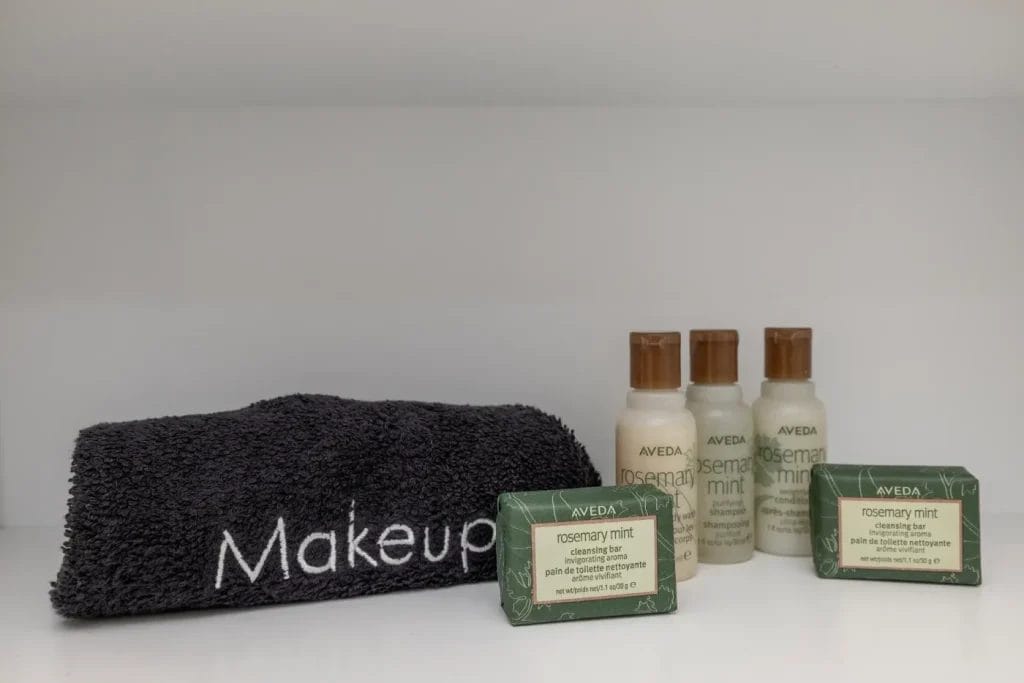 A neatly folded dark towel with "Makeup" embroidered in white sits beside three small bottles and two bars of Aveda rosemary mint cleansing products on a white shelf.