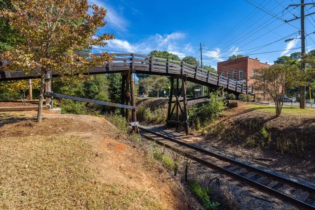 A wooden pedestrian bridge crosses over a set of railroad tracks, surrounded by trees and a brick building under a bright blue sky with scattered clouds.