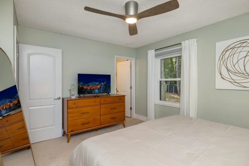 A modern bedroom with light green walls, a ceiling fan, a wooden dresser with a TV on top, a bed with a beige comforter, a tall mirror, a window with white curtains, and abstract wall art.