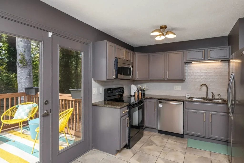 Modern kitchen with gray cabinets, stainless steel appliances, and tiled floor. A glass door opens to a deck with yellow chairs and a small turquoise table, surrounded by trees and greenery outside.
