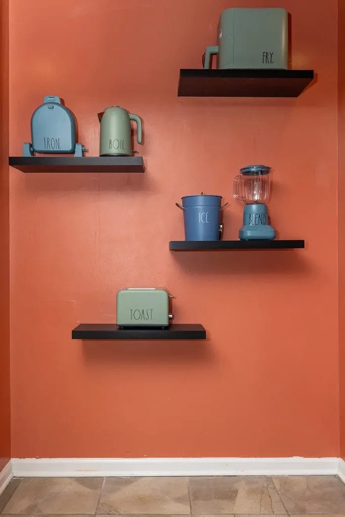 Four black floating shelves on an orange wall display pastel blue and green appliances and containers labeled “FRY,” “IRON,” “BOIL,” “ICE,” “BLEND,” and “TOAST.” The floor has beige tiles.