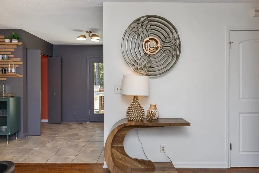 A modern entryway features a curved wooden console table with a patterned lamp, a small decorative vase, and a large circular metal wall art above. The space opens to a tiled kitchen with dark gray and orange walls.