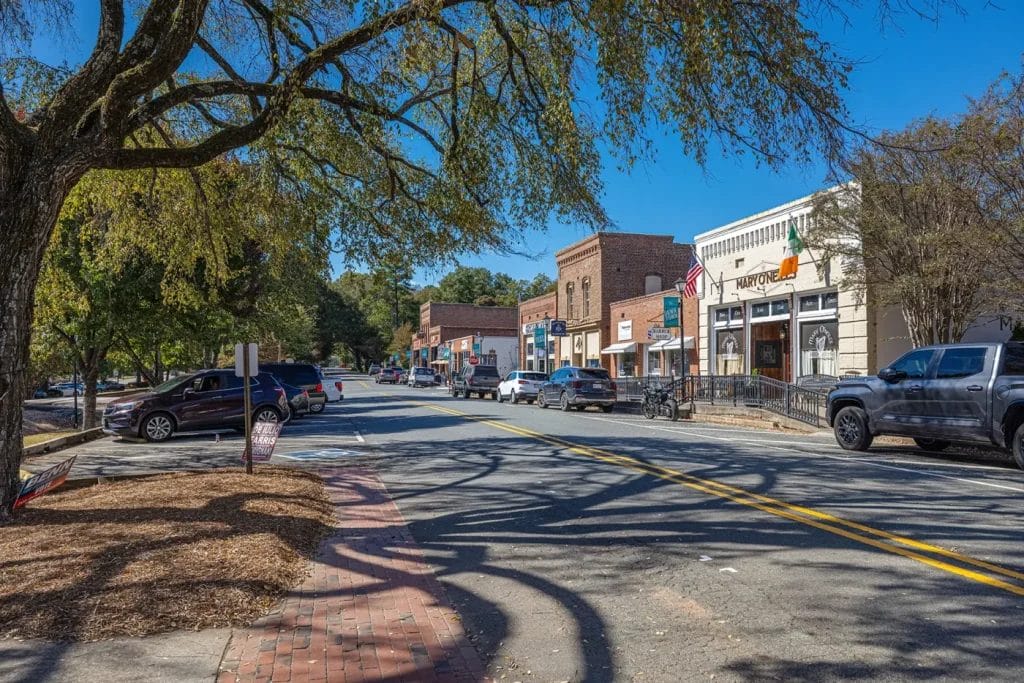 A small-town street with parked cars, brick buildings housing shops and restaurants, and trees lining the sidewalk under a clear blue sky. Shadows from branches stretch across the road.
