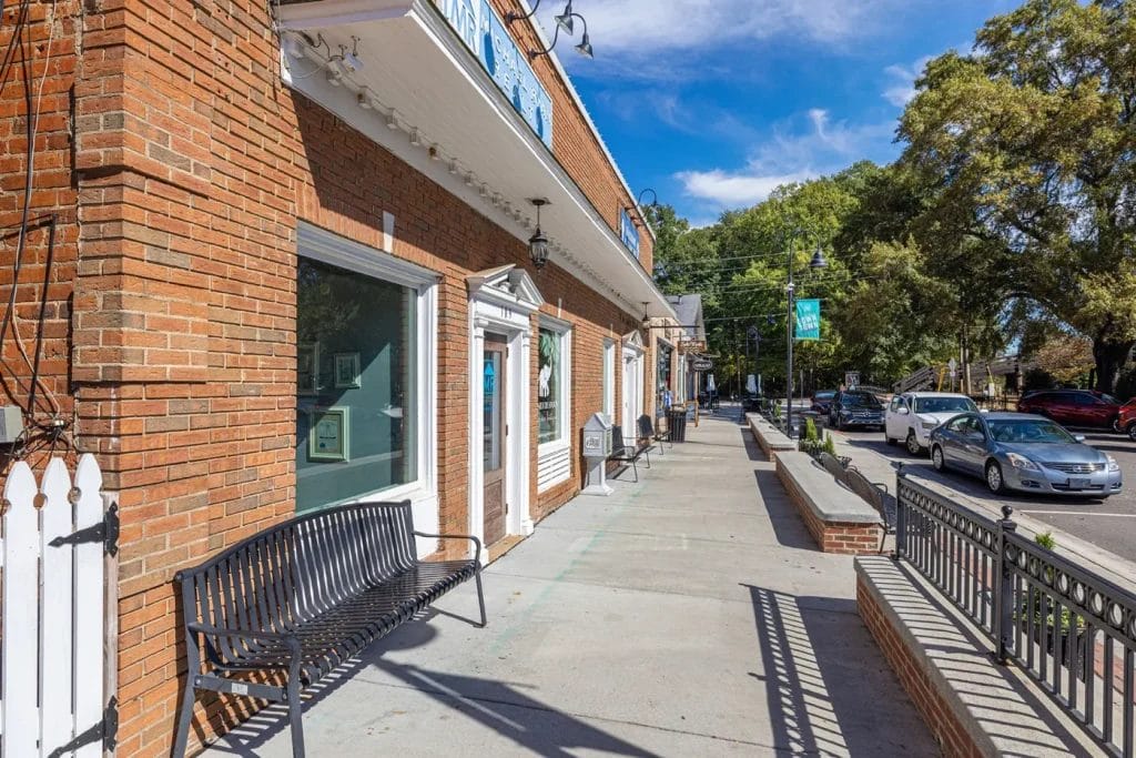 A sunny sidewalk with a black bench beside brick storefronts, trees lining the street, and several parked cars along the curb under a blue sky with scattered clouds.