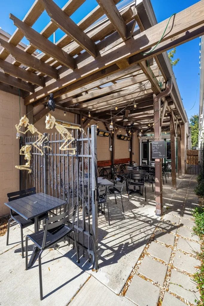 A covered outdoor patio with tables and chairs is decorated with hanging skeletons on a metal fence. String lights are draped above, and a sign hangs on a wooden post. The scene is sunny and inviting.