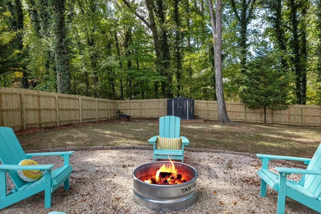 A backyard with turquoise Adirondack chairs around a metal fire pit with a bright fire, set on gravel, enclosed by a wooden fence, and surrounded by tall trees.